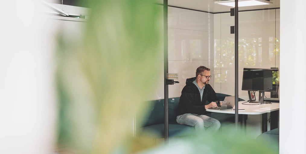Eine Person sitzt mit Laptop an einem Schreibtisch in einem Büro hinter Glaswänden; grüne Pflanzen im Vordergrund.