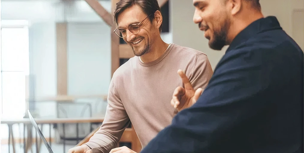 Zwei Männer arbeiten an Laptops an einem Tisch in hellem Büro; einer trägt Brille, lächelt; der andere in dunklem Hemd.
