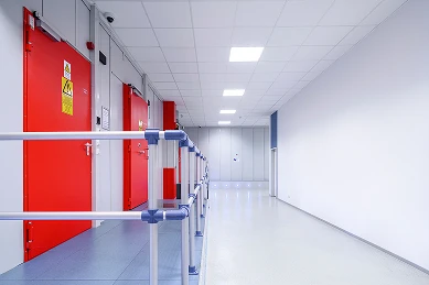 Heller Flur mit roten Türen links, blauem Geländer und weißer Decke mit Leuchten.