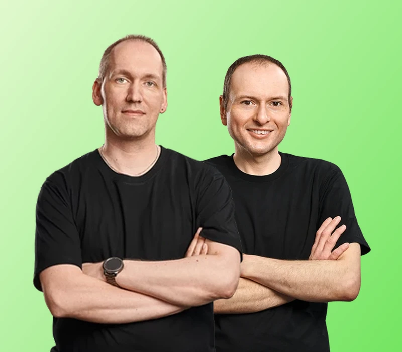 Zwei Männer in schwarzen T-Shirts stehen nebeneinander, Arme verschränkt, vor grünem Farbverlauf.
